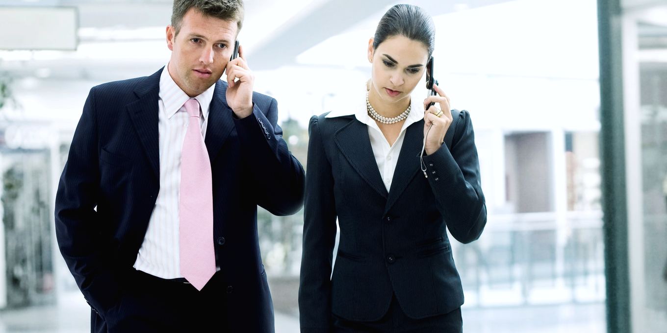 Two office workers walk through a lobby while talking on their phones